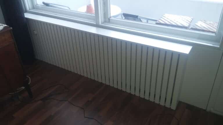 Valkoinen rimallinen patterisuoja ikkunan alla tumman puulattian vieressä.